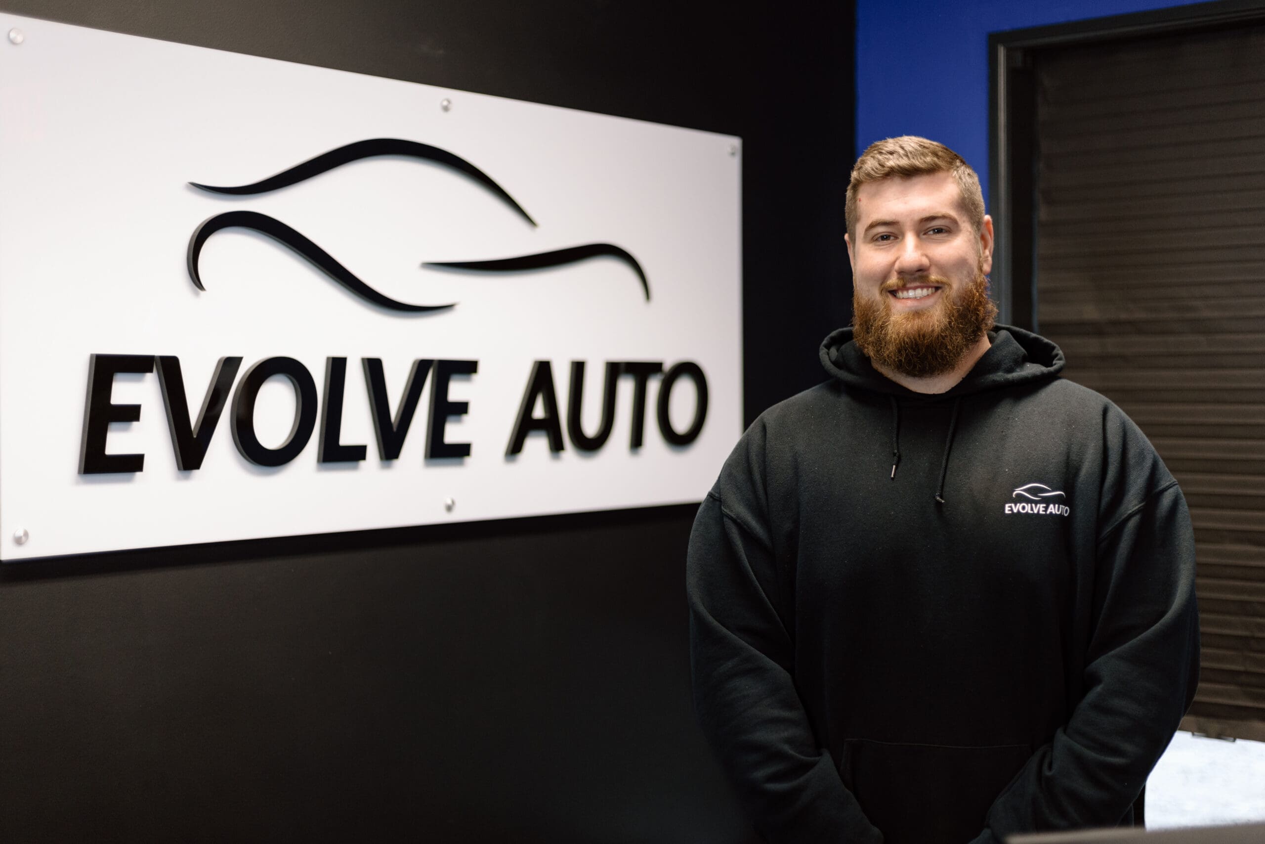 Brendan, one of Evolve Auto's Service Writers, smiles at the camera. Brendan has short ginger hair and a beard, and wears a black Evolve Auto pullover sweatshirt. Brendan is standing next to an Evolve Auto sign indoors.