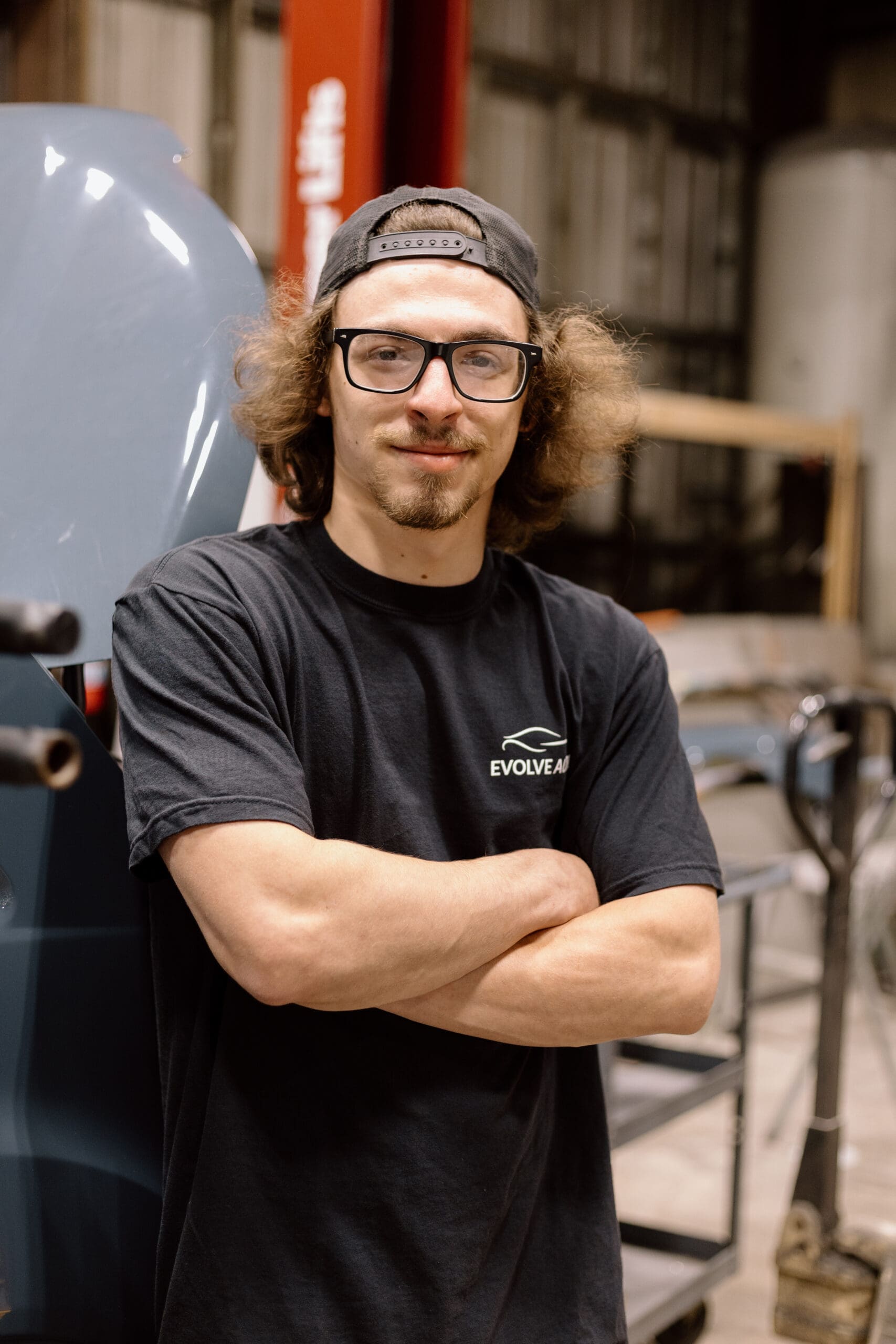 Cody, one of the Auto Body Technicians at Evolve Auto, poses with a smile and his arms crossed. Cody has brown hair, wears a backwards black cap, glasses with a black rim, and a black Evolve Auto tshirt.
