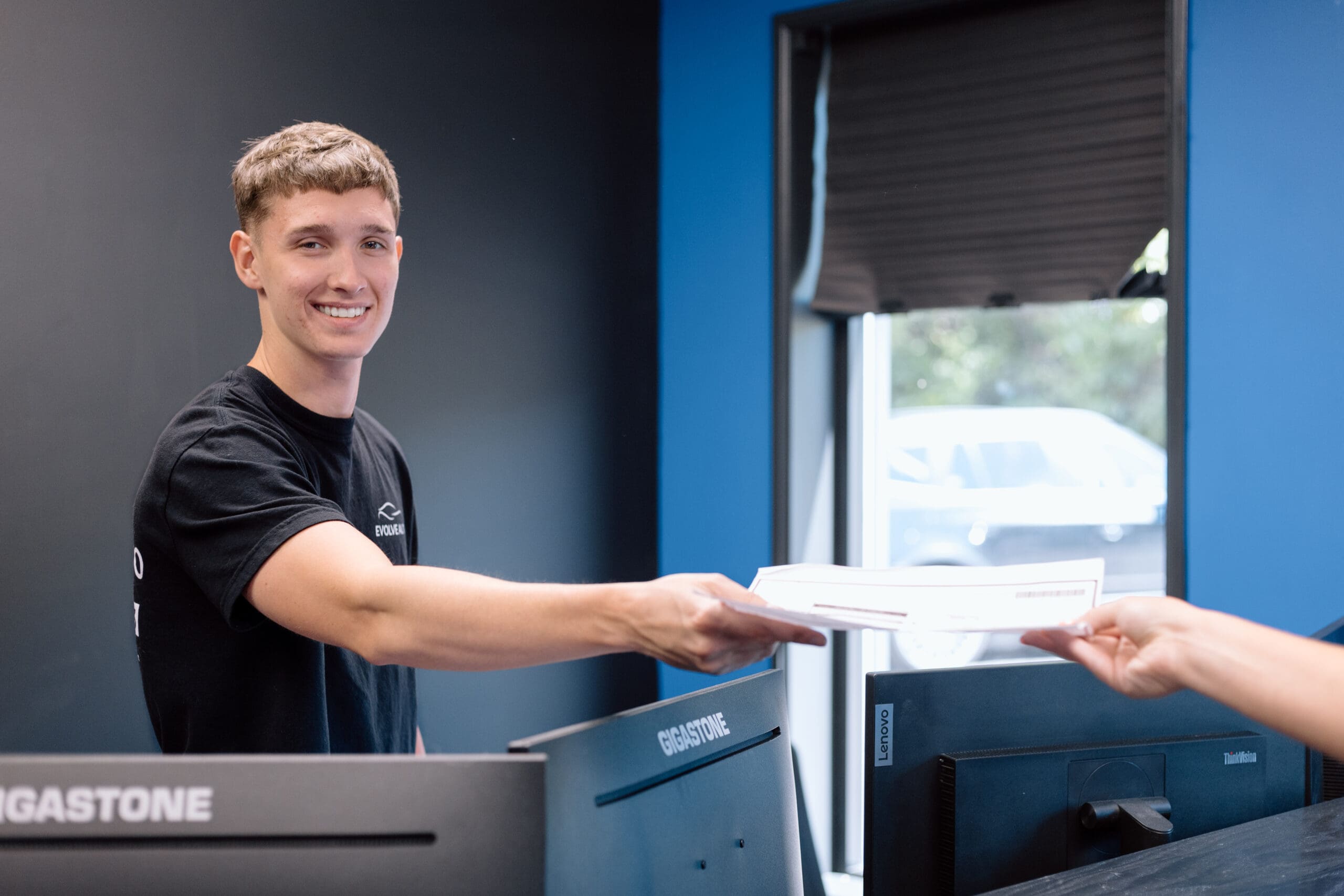 George from Evolve Auto smiles while handing a customer a certification. George has blonde hair, a black Evolve Auto shirt, and a beaming smile.