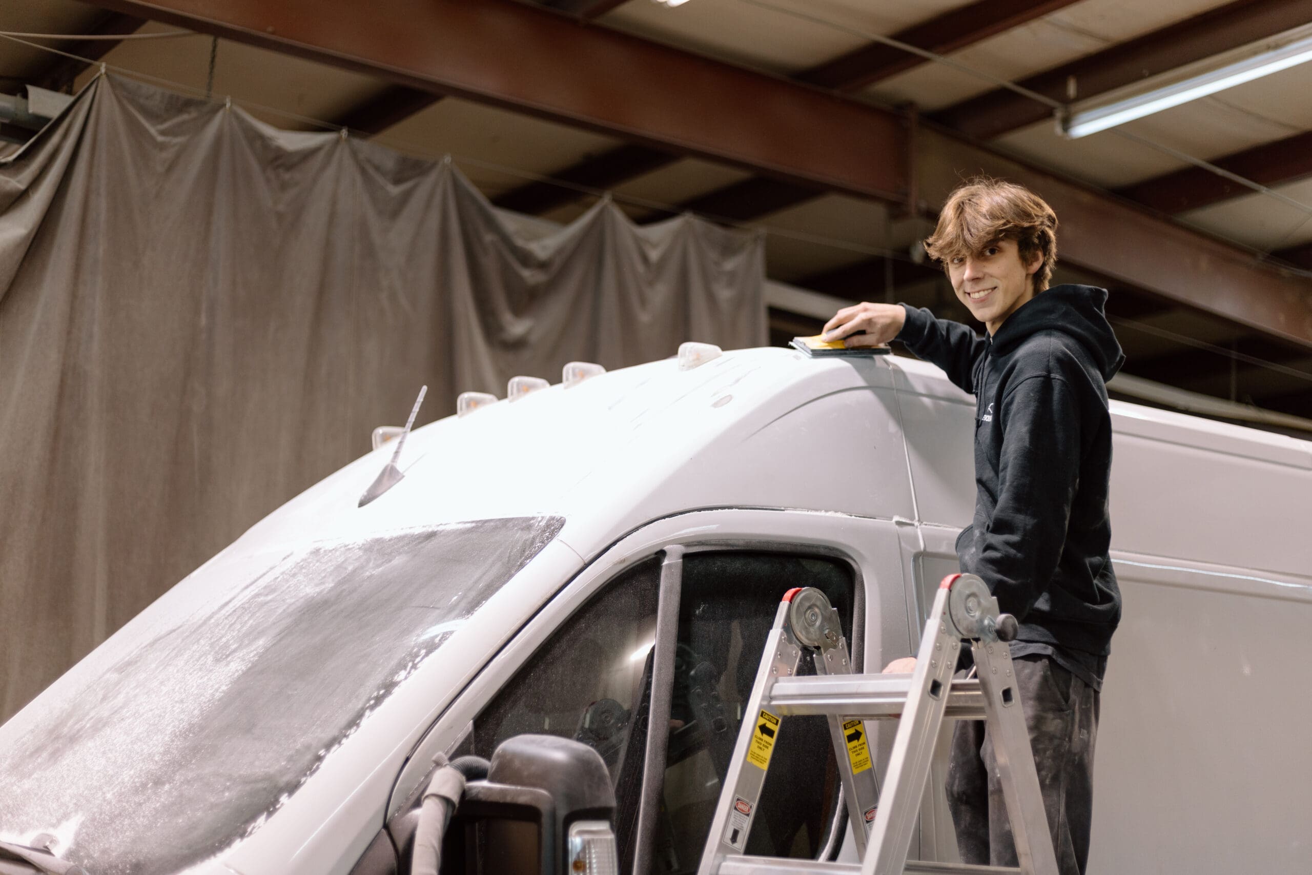 Logan, an Auto Body Technician at Evolve Auto, can be seen smiling while sanding a service van. Logan has short brown hair, brown eyes, and wears a black Evolve Auto pullover hoodie.