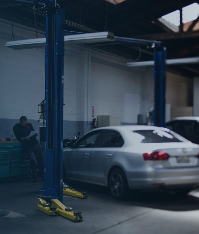 A silver sedan parked beside a two-post lift in a repair shop while a technician reviews paperwork