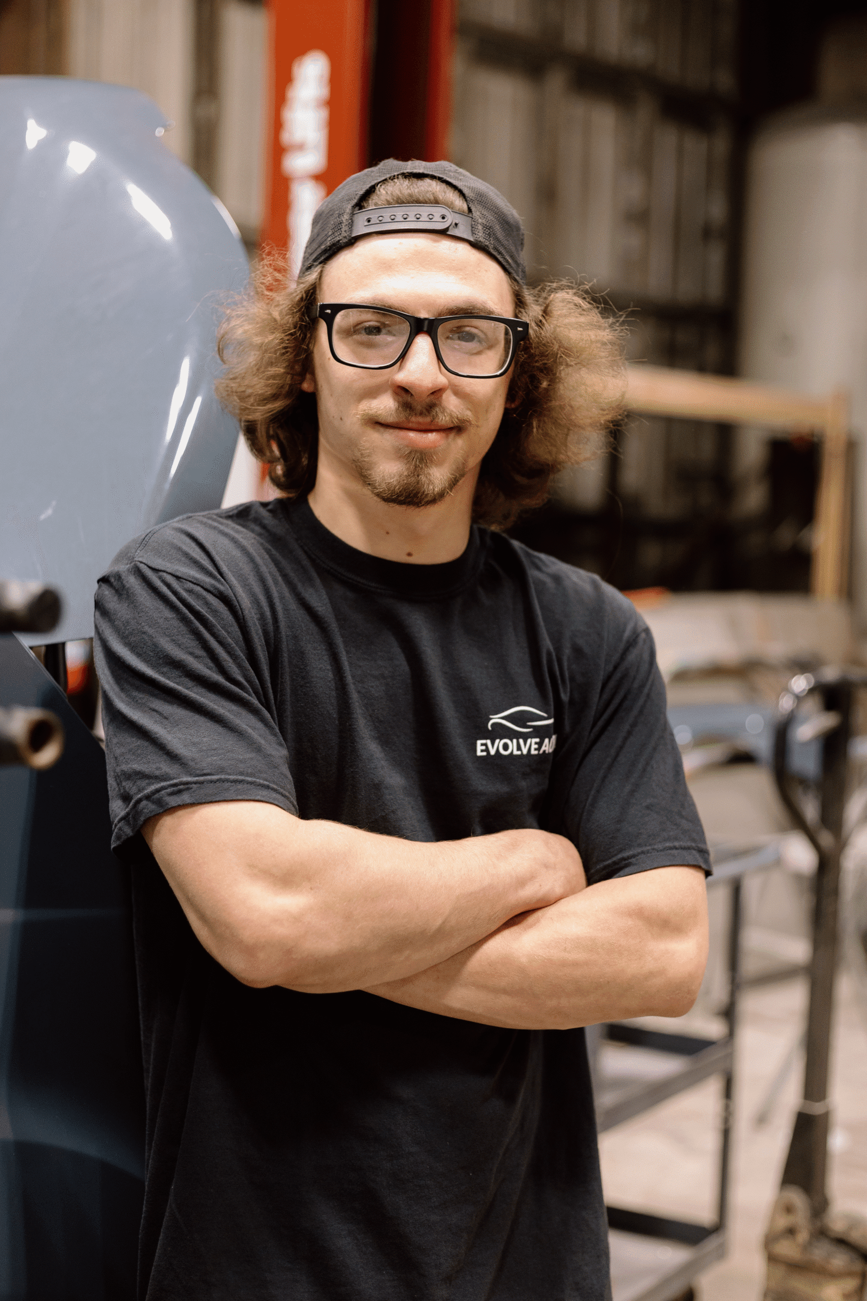 Cody, one of the Auto Body Technicians at Evolve Auto, poses with a smile and his arms crossed. Cody has brown hair, wears a backwards black cap, glasses with a black rim, and a black Evolve Auto tshirt.