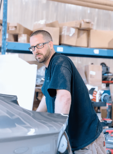 Adam, a Senior Auto Body Technician at Evolve Auto, can be seen leaning on a car. Adam has a buzz cut, a short brown beard, black rimmed glasses, and wears a black Evolve Auto tshirt.