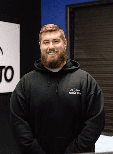 Brendan, one of Evolve Auto's Service Writers, smiles at the camera. Brendan has short ginger hair and a beard, and wears a black Evolve Auto pullover sweatshirt.