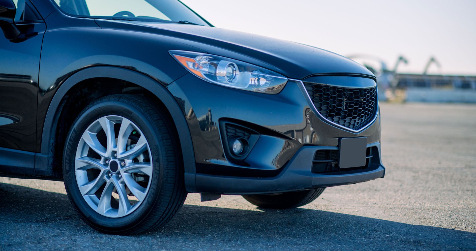 The image features the front side of black Mazda SUV Crossover. The Mazda shines underneath the sun, casting a shadow on the parking lot beneath it.