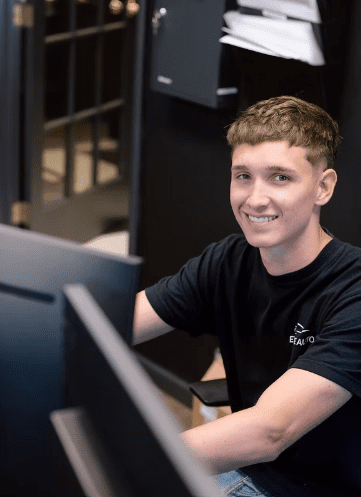 George, a Service Writer for Evolve Auto, smiles at the camera from behind a computer. George has short blonde hair, brown eyes, and wears a black Evolve Auto tshirt.