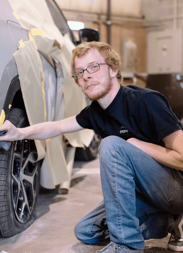 Jhonny, one of Evolve Auto's paint technicians, smiles at the camera while inspecting the wheel of a car. Jhonny has short ginger hair, a short beard, black rimmed glasses, blue jeans, and a black Evolve Auto tshirt.