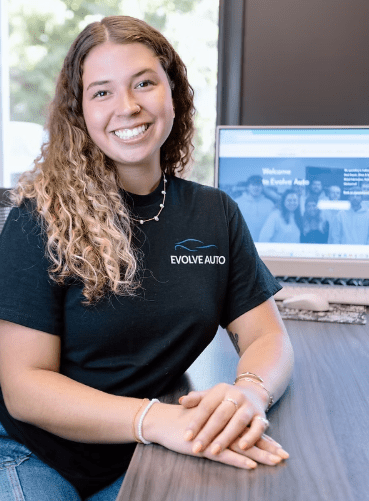 Kaci, the Marketing Manager at Evolve Auto, can be seen sitting at a desk with her hands crossed. Kaci has long curly blonde hair, a silver necklace, a black Evolve Auto tshirt, blue jeans, bracelets, and a beaming smile.