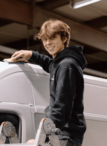 Logan, an Auto Body Technician at Evolve Auto, can be seen smiling while sanding a service van. Logan has short brown hair, brown eyes, and wears a black Evolve Auto pullover hoodie.