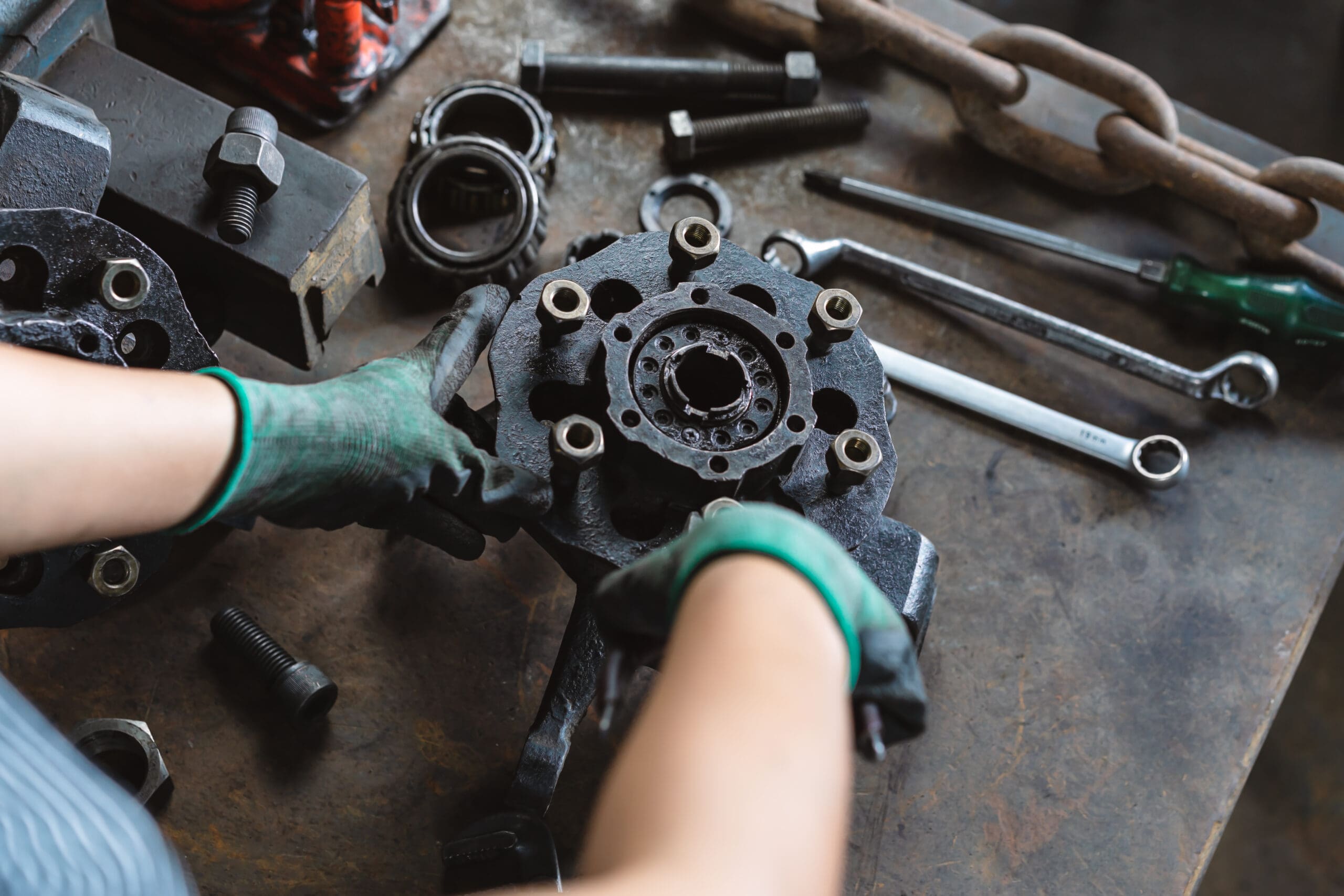 The image shows a person holding a brake caliper, a part of car brakes. The person is wearing green and black gloves, and in the background, a variety of tools can be seen scattered. Nuts, bolts, and a large chain are present on a metal table.