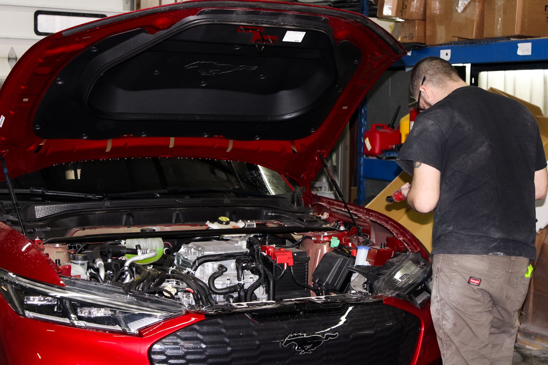 A red mustang has its hood open and is currently being serviced by Adam. Adam is to the right, inspecting a part of the car. Adam is wearing a black shirt, glasses, and brown pants.