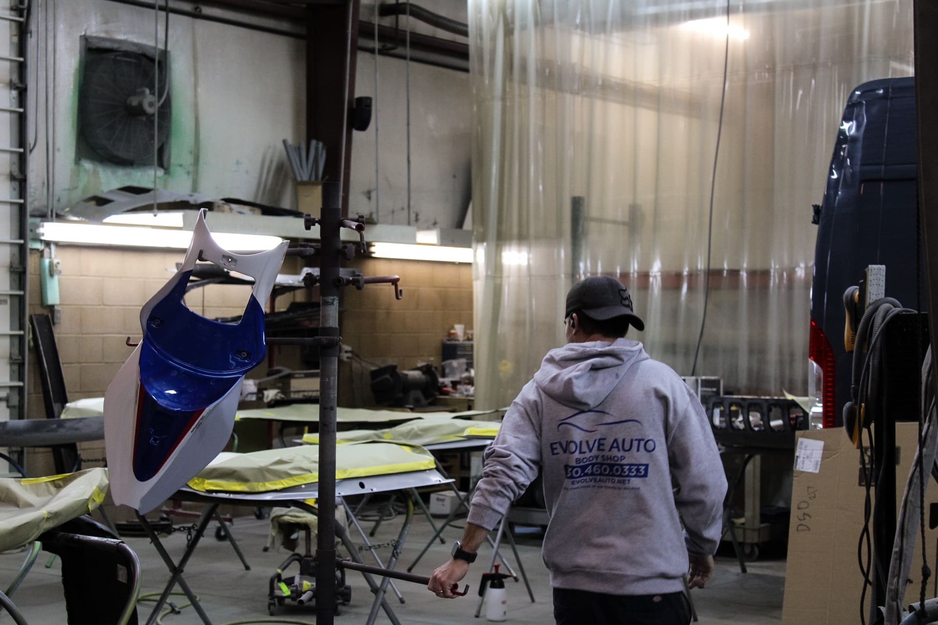 The inside of Evolve Auto's Body shop. An employee wearing a grey hoodie and a backwards cap walks by. On the left, a blue and white part for a motorcycle can be seen hanging from a rack. In the middle of the shop, car parts awaiting paint services can be seen.
