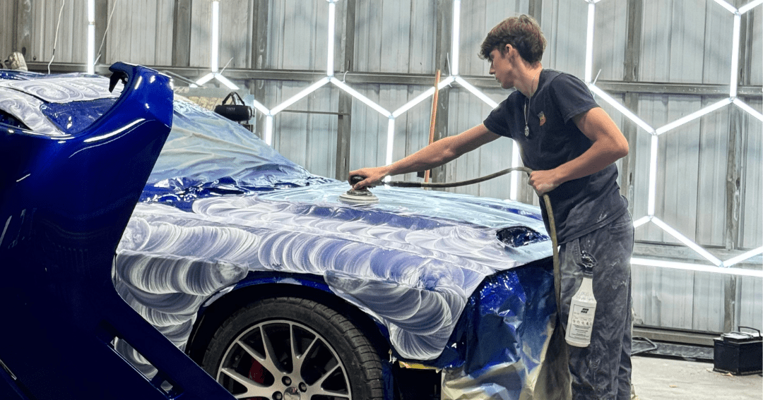 A blue Dodge Challenger receiving body services and wet sanding. An Evolve Auto employee can be seen to the right sanding the front of the vehicle, with swirled patterns all over the vehicle. The front bumper of the vehicle shows signs of damage, and to the left, a new car bumper can be seen.