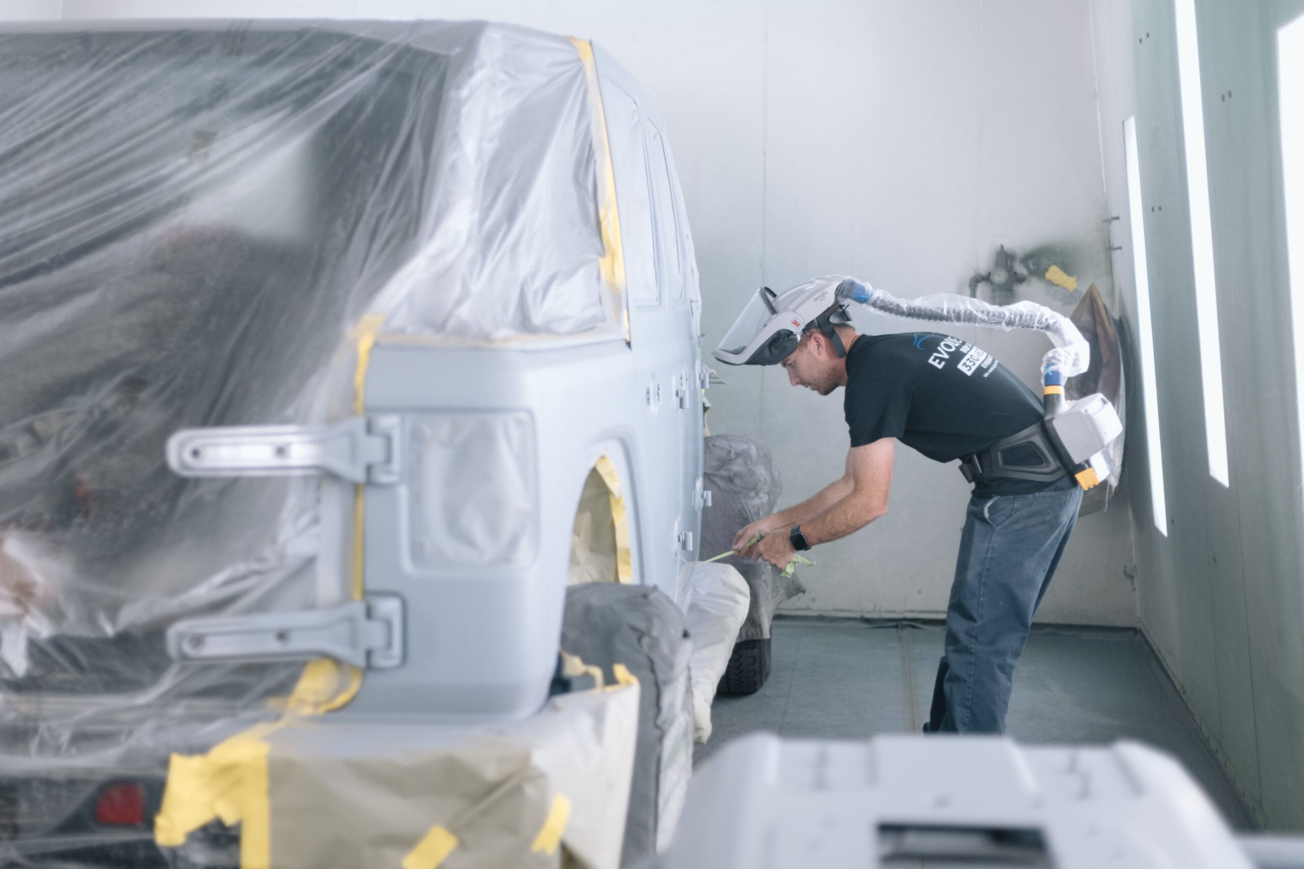 A black jeep is covered in plastic, undergoing Evolve Auto's body painting services. To the right, one of Evolve Auto's paint technicians uses a green tool to apply fresh paint to the vehicle. The technician wears protective gear, and focuses diligently on the painting process.
