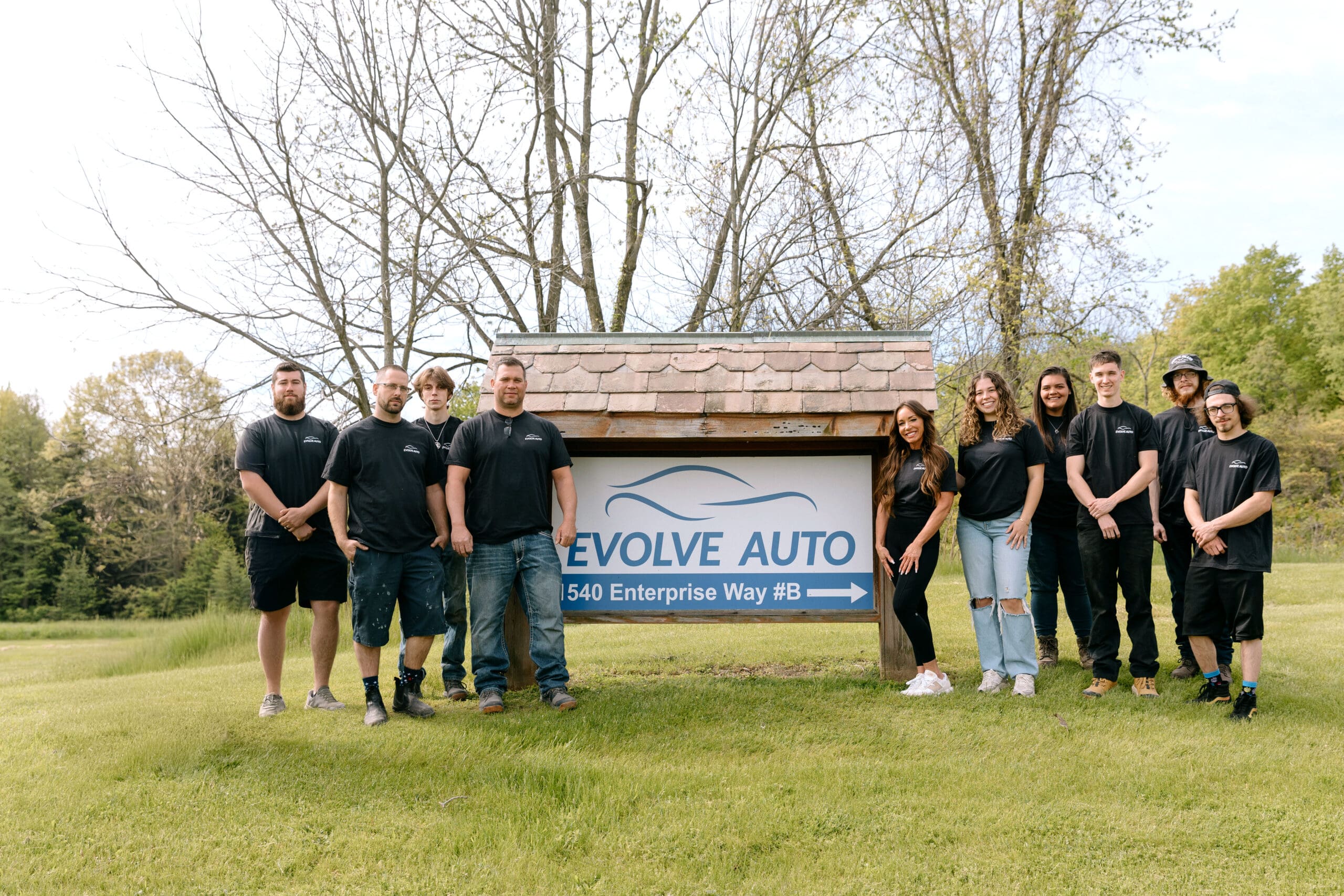 The Evolve Auto team, ten staff in black Evolve Auto shirts, standing beside the shop's sign at 1540 Enterprise Way in Kent, Ohio